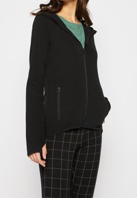 Černá mikina na zip s obepnutým střihem, dvěma bočními zipovými kapsami a měkkou, strukturovanou tkaninou. V kombinaci s černobílými kostkovanými kalhotami.