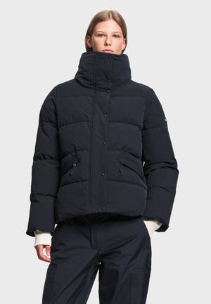 Mulher vestindo um casaco acolchoado preto com gola alta e bolsos com fecho, combinado com calças pretas, em pé contra um fundo claro e simples.