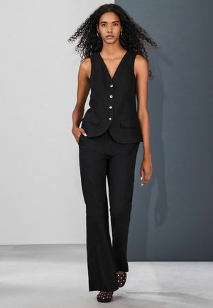 Pitkäkiharahiuksinen nainen pukeutuneena mustaan hihattomaan napilliseen liiviin ja mustiin liehuhousuihin kävelee kaksisävyistä harmaata taustaa vasten.