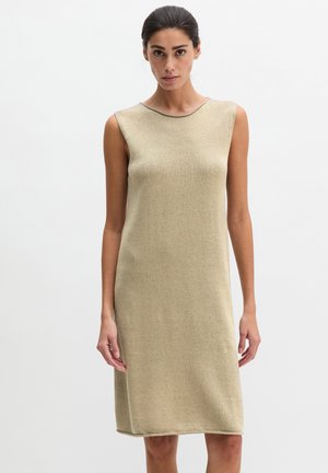Kobieta w bezrękawnym, beżowym sukienka dzianinowej sięgającej kolan, stojąca na tle jednolitego białego tła.