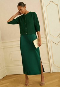 Roheline kudumiga lühikeste varrukatega kardigan, millel on nupud, kombineeritud pika rohelise satiinist kleidiga, millel on küljes lõhik, hoides kuldset käekotti.