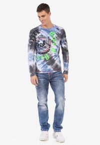 Langärmliges T-Shirt mit Batikmuster in Blau und Grau, versehen mit grafischen Drucken in Grün und Schwarz. Kombiniert mit blauen Jeans und weißen Sneakers.