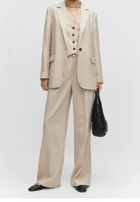 Costume beige à fines rayures avec un gilet ajusté, un blazer à un bouton et un pantalon à jambes larges. Accompagné d'un sac à main matelassé noir.