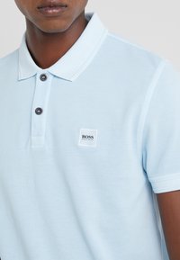 Světle modré polo tričko vyrobené z texturované tkaniny, s špičatým límečkem, dvěma tmavými knoflíky a čtvercovou nášivkou loga na hrudi.