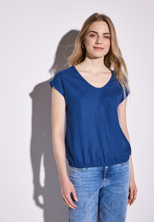 Blaues kurzärmliges Shirt mit V-Ausschnitt und elastischem Saum. Weicher, glatter Stoff mit dezentem Schimmer. Kombiniert mit hellblauen Denim-Jeans.