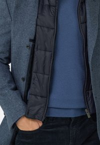 Niebieski płaszcz w jodełkę z czarną pikowaną podszewką, noszony na jednolitym niebieskim swetrze. Posiada zapięcie na guziki i boczne kieszenie. Widoczne ciemne spodnie.