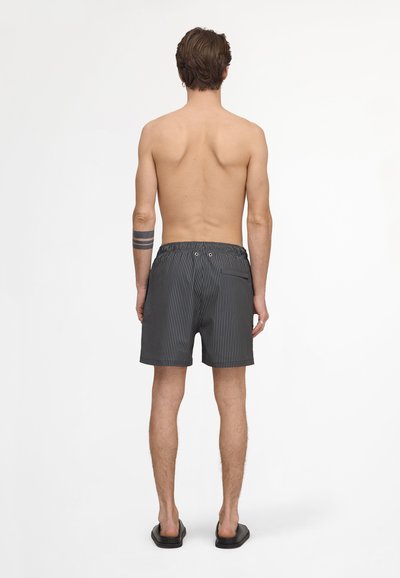 Homme torse nu, debout de dos, portant un short à fines rayures noir et blanc, des sandales noires, avec un tatouage triple bande sur l'avant-bras gauche.