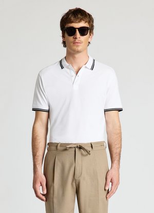 Uomo con occhiali da sole neri, polo bianca a maniche corte con bordo nero e pantaloni beige a vita alta con cintura annodata, in piedi davanti a uno sfondo grigio.