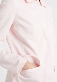 Vaaleanpunainen takki, jossa on kauluksellinen muotoilu, pitkät hihat ja sivutaskut. Kangas näyttää sileältä ja siinä on hieno kiilto.