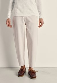 Persona con pantalones de corte estrecho color crema, una camisa de manga larga color crema y zapatos de cuero marrón con cordones, de pie sobre un suelo liso.