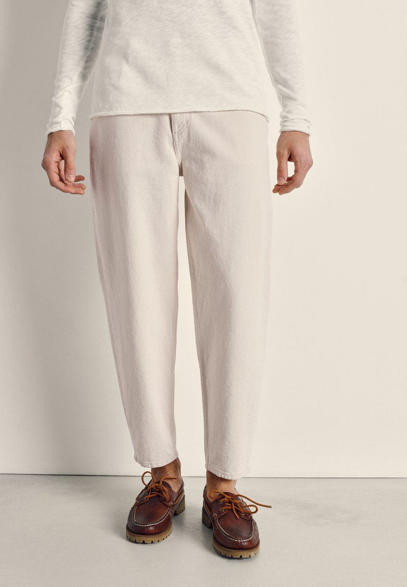 Persona con pantalones de corte estrecho color crema, una camisa de manga larga color crema y zapatos de cuero marrón con cordones, de pie sobre un suelo liso.