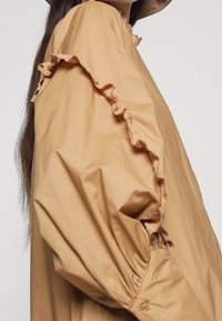 Chemise en coton couleur beige avec des accents à volants le long des manches, une coupe décontractée et une texture lisse, mettant en avant des coutures détaillées.