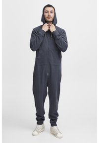 Tamnoplavi zip-up kombinezon s kapuljačom, od mekanog materijala. Ima prednje džepove i elastične manžete. Model nosi bijele teniske.