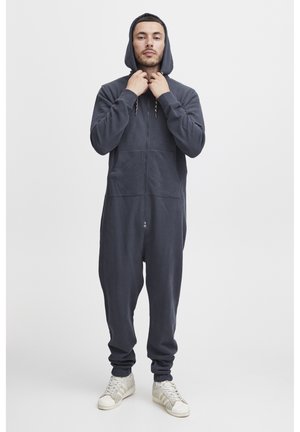 Mörkblå zip-up jumpsuit med huva, tillverkad av mjukt tyg. Har framfickor och elastiska ärmslut. Modellen bär vita sneakers.