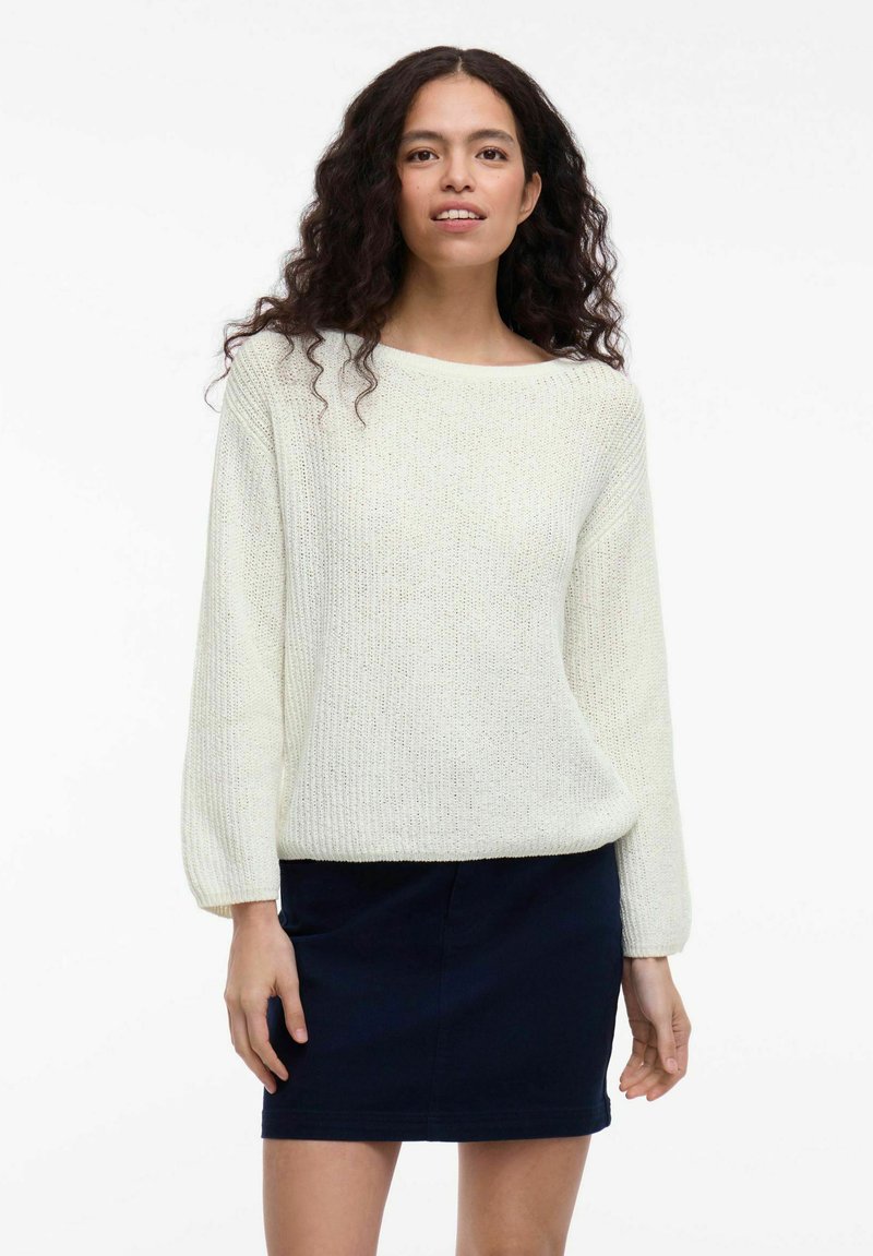 Jasna kremowa swetrowa bluzka z szerokim dekoltem w kształcie łódki i długimi rękawami, zestawiona z ciemnonawowym mini spódnicą. Teksturowany materiał.