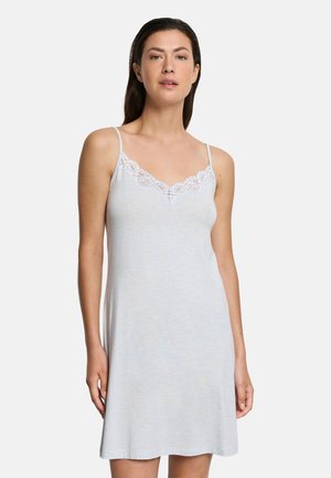 Kvinde iført lysegrå ærmeløs kjole med blondekant ved halsudskæringen, stående mod en ensfarvet hvid baggrund.