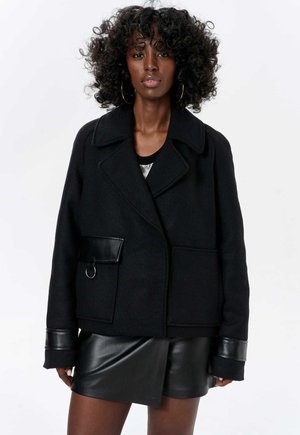 Black wool blend jacket with a large collar, leather accents, front pocket, and a round metal ring detail. Worn over a black leather mini skirt.