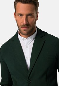 Blazer verde scuro con rever a punta, chiusura con un bottone e taschino sul petto. Indossato sopra una camicia bianca fresca con dettagli sui bottoni.