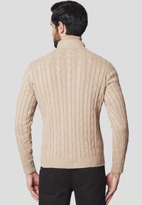 Beżowy sweter z splotem w warkocze z wysokim kołnierzem, żebrowanymi mankietami i dołem. Ma teksturowane wzory na całej tkaninie dla dodatkowego efektu.