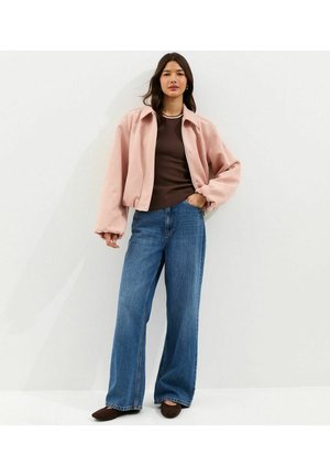 Vrouw staand, gekleed in een lichtroze oversized jas, een bruin geribd shirt, brede blauwe jeans en bruine schoenen tegen een effen lichte achtergrond.