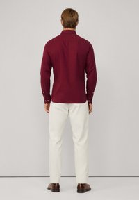 Camicia a maniche lunghe color bordeaux con colletto a punta e pantaloni bianchi; presenta polsini con bottone e una vestibilità sartoriale, vista dalla parte posteriore.
