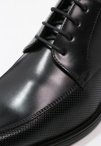 Chaussure habillée en cuir noir à finition brillante, présentant un motif perforé sur le côté, des lacets droits et un design élégant.