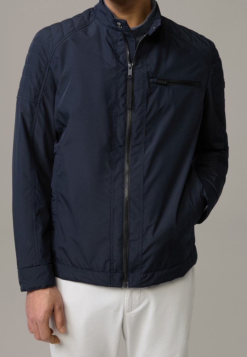 Personne portant une veste bleu foncé zippée avec des épaules matelassées et un pantalon blanc, une main dans la poche, fond neutre.