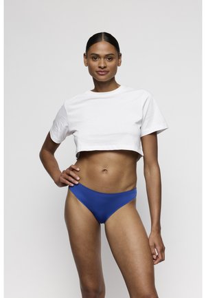 Vrouw met donker haar in een wit cropped t-shirt en blauwe bikinibroekjes staande met hand in de heup tegen een lichtgrijze achtergrond.