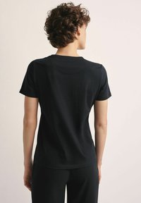 Osoba z krótkimi kręconymi włosami, ubrana w prostą czarną koszulę z krótkim rękawem i czarne spodnie, pokazana od tyłu na neutralnym tle.