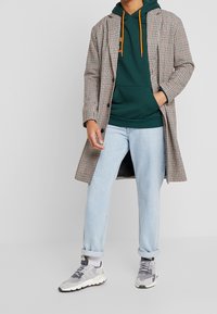 Ternet frakke med langt snit, grøn hættetrøje med orange snørrebånd, lyseblå jeans og grå sneakers. Har kontrasterende teksturer og farver.