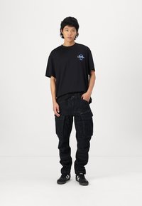 Black short-sleeve T-shirt with blue graphic detail, paired with dark cargo pants featuring multiple pockets and zippers. Dark sneakers complete the look.