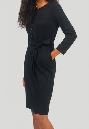 Schwarzes Kleid mit langen Ärmeln, Knopfleiste vorne, Taillenband und seitlichen Taschen. Glatte Textur und knielanges Design. Schlichter, vielseitiger Stil.