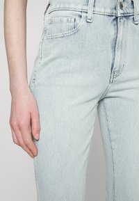 Jeans de ganga azul claro com um ajuste slim, apresentando bolsos frontais, fecho de zip e um leve desbotamento na textura do tecido.