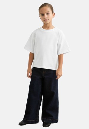 Fehér, túlméretezett, rövid ujjú póló, széles szárú sötétkék farmerral párosítva. A farmer narancssárga varrással és lazább szabással rendelkezik.