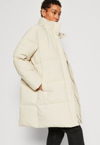 Veste matelassée beige avec col montant, texture quiltée et poches latérales. Fermeture à glissière et coupe décontractée, fabriquée en matériau léger.