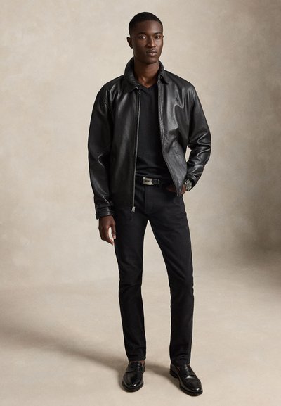 Moški stoji, oblečen v črno usnjeno jakno, črno majico z V-izrezom, črne hlače, črni pas, črne elegantne čevlje in ročno uro na bež ozadju.