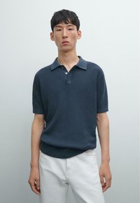 Polo shirt blu navy a maglia con maniche corte, dotato di colletto e patta con tre bottoni, abbinato a pantaloni bianchi.