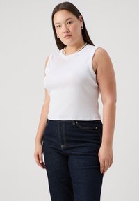 Top corto sin mangas blanco en tejido acanalado, combinado con jeans de tiro alto en azul oscuro que presentan costuras visibles y bolsillos frontales.