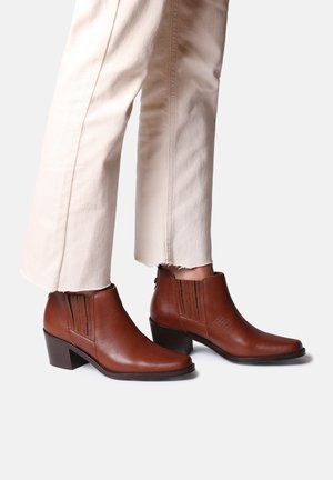 Brown leather ankle boots with a square toe, textured finish, side elastic accents, and a low block heel. Worn with light-colored pants.