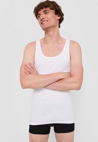 Vit bomullstopp med rund halsringning, tillsammans med svarta boxershorts, bärs av en person med ljus hy och kort lockigt hår.