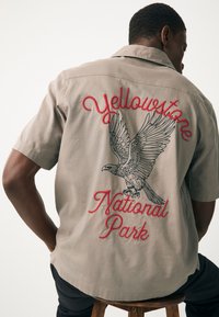 Chemise beige à manches courtes avec une broderie détaillée d'aigle et le texte « Yellowstone National Park » en rouge, avec une coupe décontractée et une texture douce.
