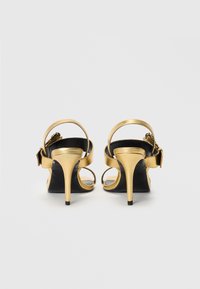 Gold high-heel sandals with a sleek metallic finish, open-toe design, adjustable ankle straps, and a stiletto heel. Black sole visible.