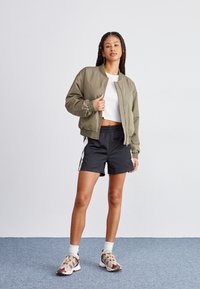 Chaqueta bomber verde olivo con puños elásticos, combinada con pantalones cortos deportivos negros con rayas laterales blancas. Camiseta blanca y zapatillas multicolores.