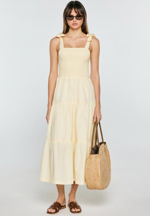 Kobieta ubrana w jasnopomarańczową, bezrękawnikową, wielowarstwową sukienkę midi, okulary przeciwsłoneczne, brązowe sandały i trzymająca dużą plecioną słomianą torbę.