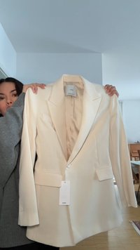 Femme tenant un blazer couleur crème avec des revers et des poches avant dans une pièce lumineuse avec un sol en bois et des meubles.