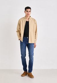 Camisa beige de botones sobre una camiseta de tirantes negra, combinada con jeans azules y zapatillas marrones. Diseño simple con un corte relajado.