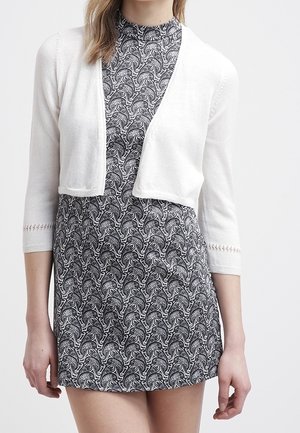 Vrouw draagt een zwart-witte jurk met een paisley-patroon en een wit cropped vest met driekwart mouwen.