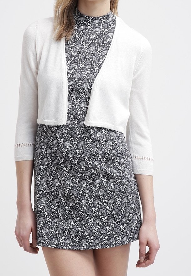 Femme portant une robe à motif cachemire noir et blanc avec un cardigan court blanc aux manches trois-quarts.