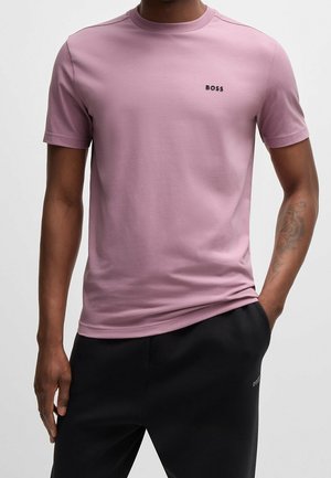 Homme portant un t-shirt moulant rose poudré à manches courtes avec le logo "BOSS" sur la poitrine et un pantalon noir, une main dans la poche, sur un fond uni.