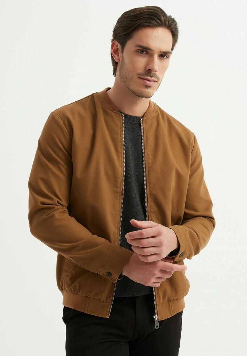 Veste bombers marron avec fermeture éclair à l'avant, poignets côtelés et taille côtelée. Portée par-dessus une chemise gris foncé, elle présente une texture lisse et un design minimaliste.